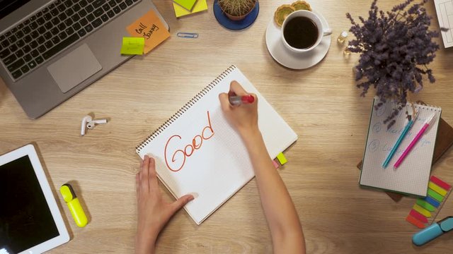 Girl writes on a piece of paper a phrase - "Good Day To Start", sitting at work table, first-person view of hands.