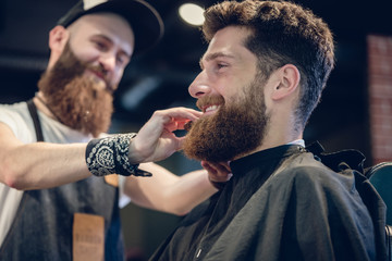 Close-up of the hands of a skilled barber using a handless brush with boar bristles, while grooming the beard of his young customer in a trendy hair salon for men only