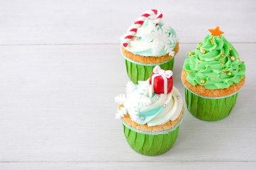 New year or christmas green cupcakes with whipped cream, decorated with a christmas tree, candy cane, a snowflake, a red box, silver and gold confectionery balls on white wood table.