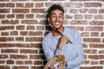 Young African-American artist with palette and brush against brick wall