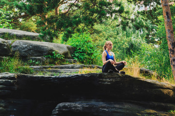 Blonde woman meditates in easy pose outdoors on a rock in the forest. Yoga nature concept.