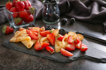 Slate board with tasty thin pancakes and berries on dark table