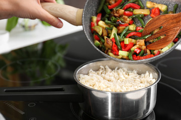 Woman adding vegetables into saucepan with boiled rice in kitchen