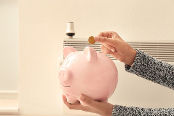 Woman putting coin into piggy bank near radiator at home. Heating saving concept
