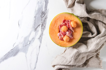 Melon and ham or prosciutto salad served in half of Cantaloupe melon on linen cloth over white marble background. Flat lay, space