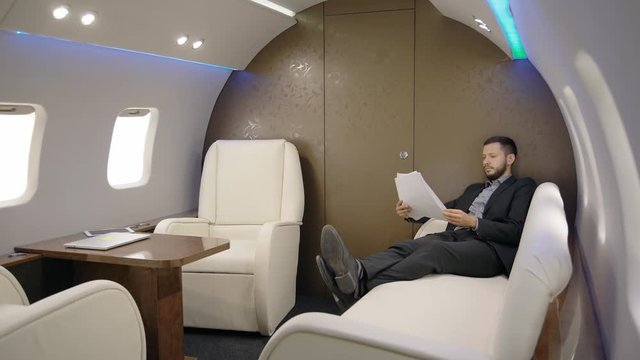 Young investor expert analyst respectable man is working on project sitting in private airplane, handsome businessman is looking at documents during business traveling, resting on leather seats