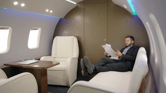Business owner chief banker director is reading contract on leather sofa of airplane, stylish bearded businessman working with documents during flight, having time in luxurious interior. Concept