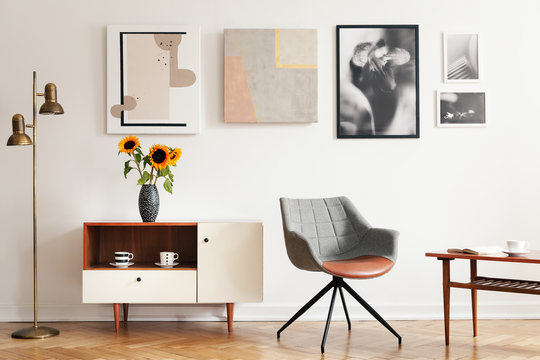 Grey armchair next to white cupboard with sunflowers in apartment interior with posters. Real photo