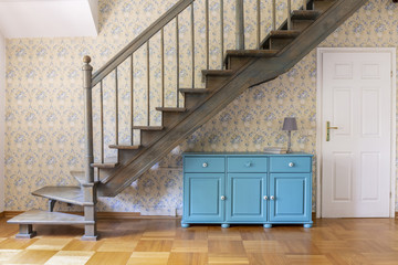 A light blue three door cabinet standing under gray staircase against a wall with flower wallpaper...