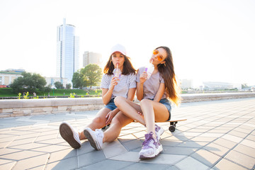 Two Young Woman sitting on Skateboard Happy Smiling. Playful Friends Enjoy Sunny day. Outdoor Urban. Beautiful Model Girl in Fashion Trendy Outfit