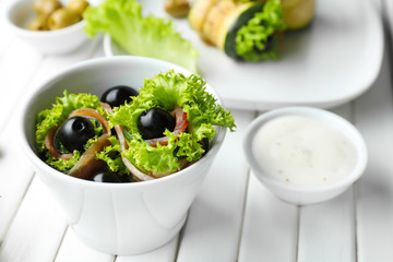 Tasty zucchini rolls with prosciutto and olives in bowl on white table