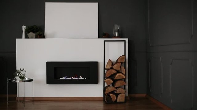 Frame with wood placed next to eco fireplace in the video of dark grey living room interior with wainscoting on wall, fresh plants and mockup poster on shelf. Fire burning in the fireplace