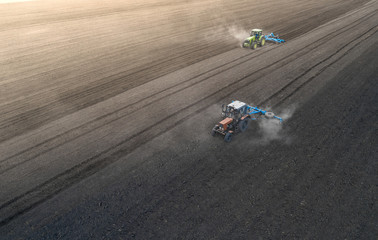 Aerial view of two blue tractors plows the earth in field on a summer day against a black earth...