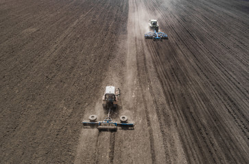 Aerial view of two blue tractors plows the earth in field on a summer day against a black earth...