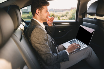 Portrait of handsome man in suit working on laptop, while back sitting in business class car