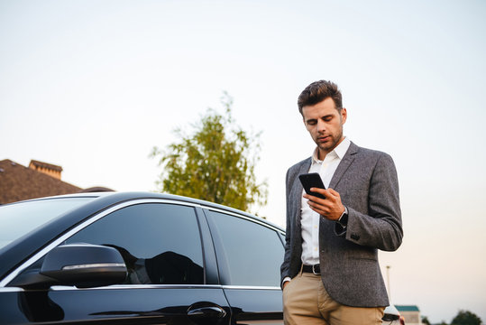 Portrait of rich businessman wearing suit, standing near his luxury black car, and using smartphone while holding in hand