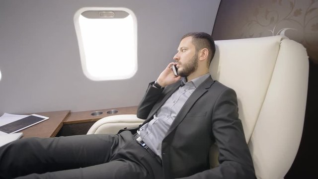 Young bearded investor expert analyst businessman discussing business during flight, sitting in private plane, man talking on phone, having time in leather armchair, looking out window. Concept: vip