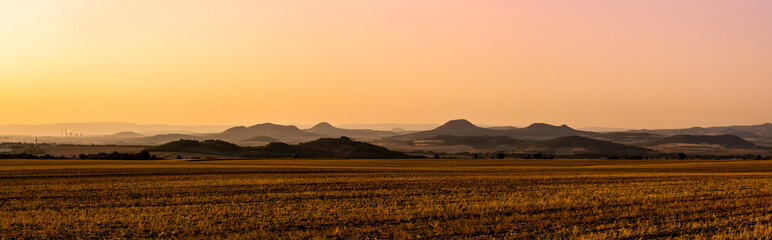 Panorama of Central Bohemian Uplands (Ceske stredohori) during sunset with pink and orange color tint on a golden harvested field, town and factory in the background