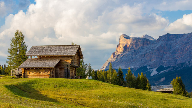 Wooden cottage on a green grass meadow with forest, valley, high sharp mountains and cloudy blue sky in the background
