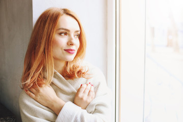 Fototapeta premium Beautiful redhead young woman with red lipstick on lips sitting by the window of coffee shop, wearing winter autumn season white knitted sweater & big loose scarf on shoulders. Background, close up.