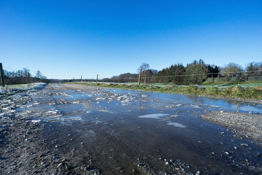 Low angle photo of puddle with ice on dird road with electric fence on the sides