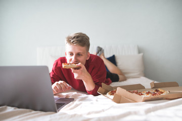 Young happy man is lying on the bed with a box of pizza, uses a laptop and eats a piece of pizza.Teen is lying on the bed, watching movies and eating delivery pizza. - obrazy, fototapety, plakaty