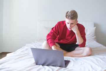 Portrait of a concentrated young man sitting on a bed in a bright room with a notebook and working. Serious freelancer is sitting on the bed in a bright, cozy room and working on a laptop.