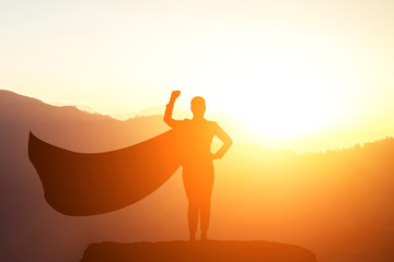 silhouette of a business woman superhero with a cloak standing on top of a mountain in a pose of success in the sunlight
