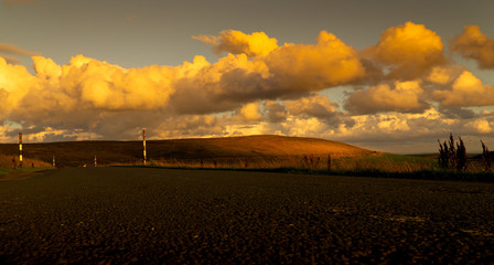 A tarmac road leads to the distance, as light through the clouds highlights a section of farmland in a moorland landscape scene as the sun sets.  The colours are autumnal with an orange glow. 