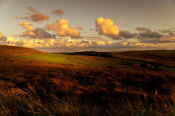 Light through the clouds highlights a section of farmland in a moorland landscape scene as the sun sets.  The colours are autumnal with an orange glow. 