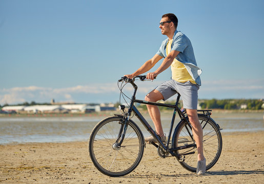 people, leisure and lifestyle concept - happy young man riding bicycle along summer beach