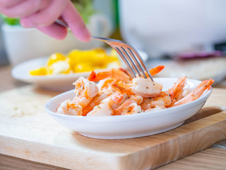 Woman 's hand with fork, shrimps, salad in white bowl, yellow tomatoes on cutting board.