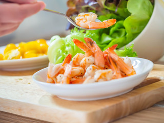 Woman 's hand with spoon, shrimps, salad in white bowl, yellow tomatoes on cutting board.