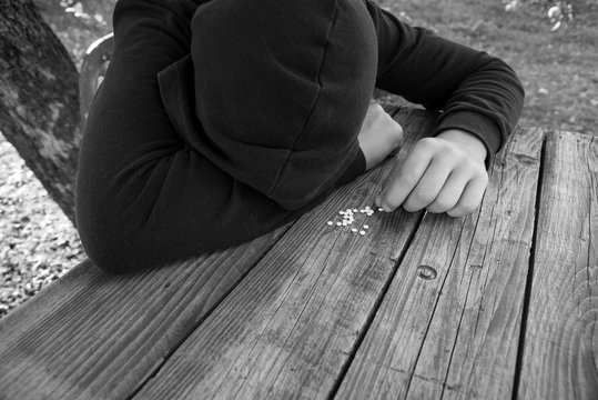 Monochrome image of a young boy wearing hood holding drugs on scratched old pine table, conceptual image of drug prevention, focus on the boys hand.