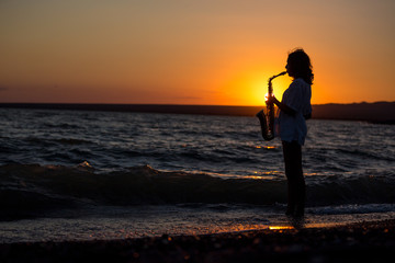 Silhouette of young sexy woman playing saxophone on the beach at sunset