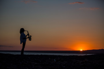 Silhouette of young sexy woman playing saxophone on the beach at sunset
