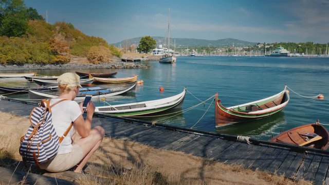 A woman with a backpack sits on the waterfront of the city of Oslo in Norway near the fishing boats. uses a smartphone. Travel around the Scandinavian countries