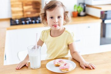 Little girl eating donuts and drinking milk on the white kitchen at home. Child is having fun with donuts. Tasty food for kids.