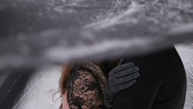 Bride in a black dress and a black veil hugging her husband in the winter in heavy snowfall. A black veil covers the head of the bride. Close-up. Gothic wedding.