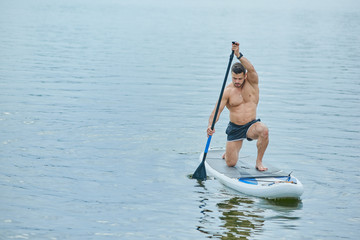 Young sportsman keeping long oar in vertical position, swimming on sup board in city lake.