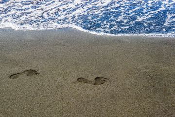 traces of human feet on black sand near the water on the beach. Background texture.