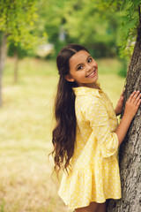 Naklejka premium Little smiling lady posing next to tree. Gorgeous petite girl wearing bright summery yellow polka dot dress looking over her shoulder smiling.