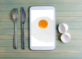 Fried egg on the tablet screen. Cutlery - knife and fork, empty eggshell. Wooden background.