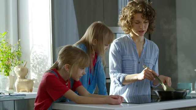 Medium shot of young curly haired woman cooking food in kitchen and talking to two daughters, who are watching