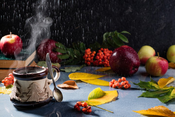 Autumn composition with rain. Steaming coffee cup, autumn leaves, rowan, apples and books on rainy day background. Autumn background with cozy atmosphere