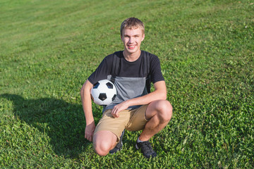 Happy teenager kneeling on the grass while holding a soccer ball.