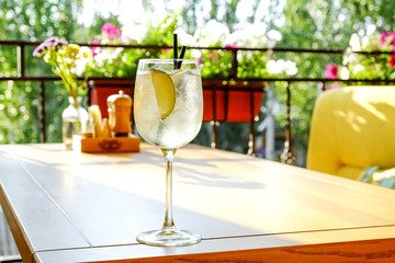 Glass of fresh summer non alcoholic refreshing cocktail with ice & lemon, standing on table of patio outside hip restaurant with city greenery view. Soft sunlight ray. Background, copy space, close up
