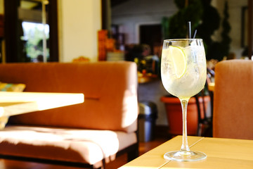 Glass of fresh summer non alcoholic refreshing cocktail with ice & lemon, standing on table of patio outside hip restaurant with cushioned seats. Soft sunlight ray. Background, copy space, close up
