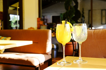 Two glasses of fresh summer non alcoholic refreshing cocktail, ice, orange & lemon on table of patio outside hip restaurant with cushioned seats. Soft sunlight ray. Background, copy space, close up
