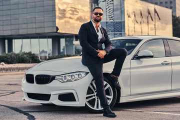 Handsome bearded male in sunglasses dressed in a black suit sitting on luxury car against a...